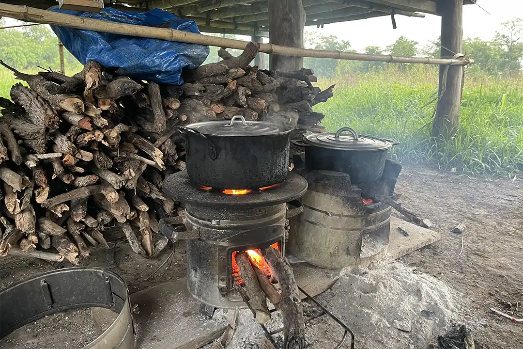 A couple of high efficiency cookstoves with pots on top. They are lit and burning well, with a gathering of wood piled behind them to keep them going.